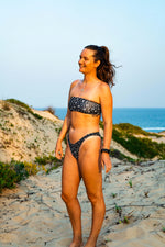 Load image into Gallery viewer, Woman in a black and white patterned bikini standing on a sandy beach with ocean and sky in the background.
