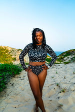Load image into Gallery viewer, Woman in a black and white patterned swimsuit standing on a sandy beach with ocean view.
