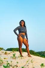 Load image into Gallery viewer, Woman in a black and white patterned swimsuit standing on a sandy beach with a clear blue sky.

