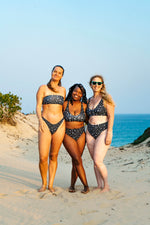 Load image into Gallery viewer, Three women in swimsuits standing on a sandy beach with ocean in the background
