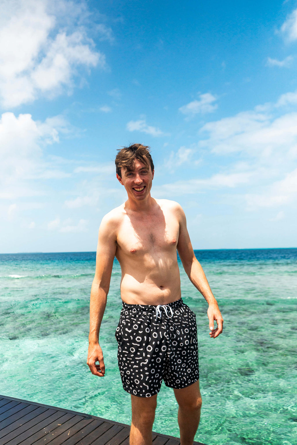 Man in swim shorts standing on a wooden deck with clear blue water and sky in the background