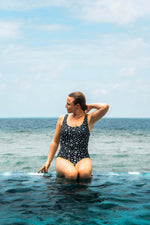 Load image into Gallery viewer, Woman in a black patterned swimsuit standing in the ocean with a clear blue sky.
