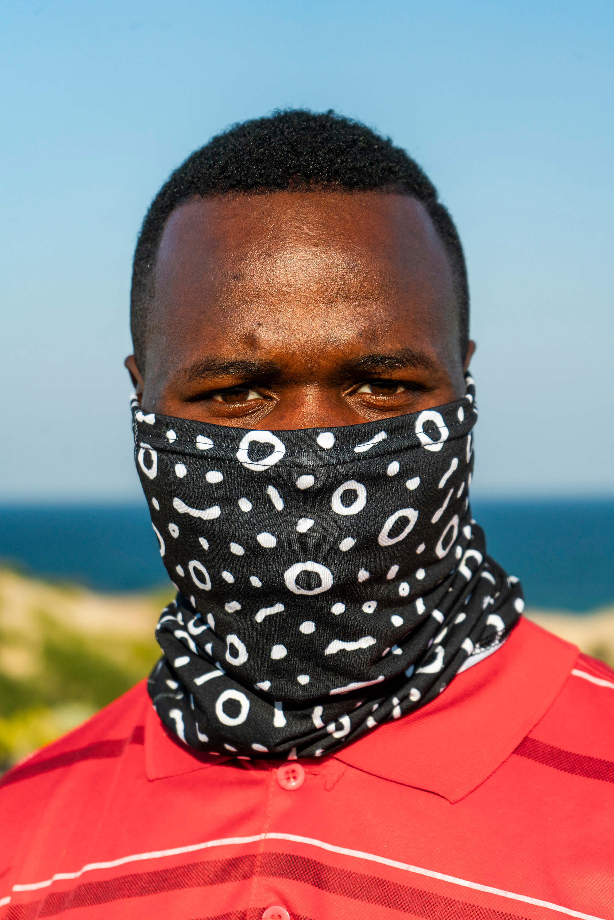Man wearing a black patterned face cover with ocean and sky in the background