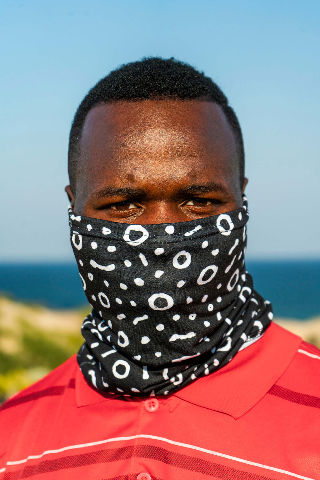 Man wearing a black patterned face cover with ocean and sky in the background