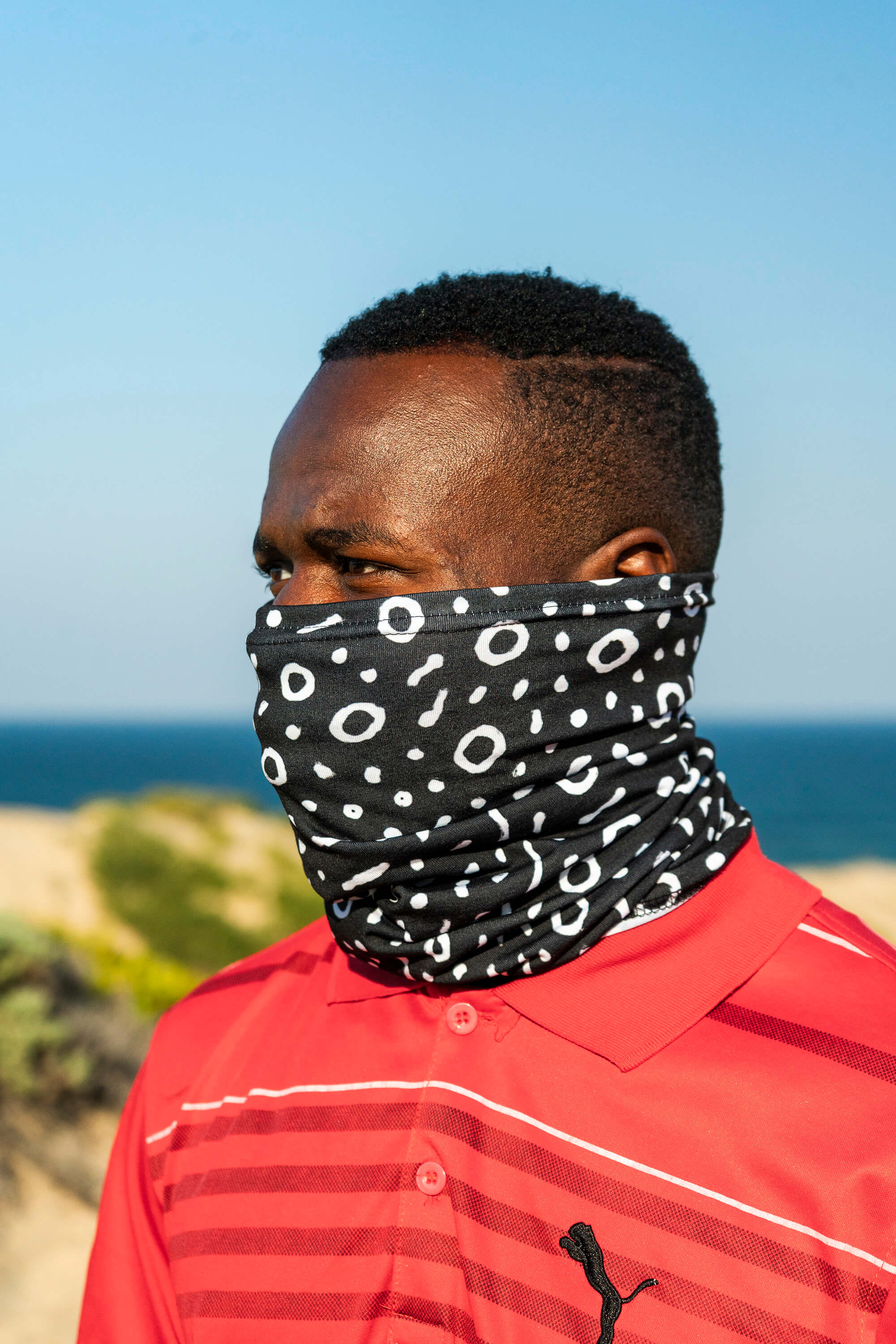 Man wearing a black and white patterned face cover in an outdoor setting with blue sky and ocean.