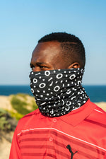 Load image into Gallery viewer, Man wearing a black and white patterned face cover in an outdoor setting with blue sky and ocean.
