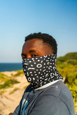 Load image into Gallery viewer, Person wearing a black patterned face cover outdoors with a clear blue sky and greenery.
