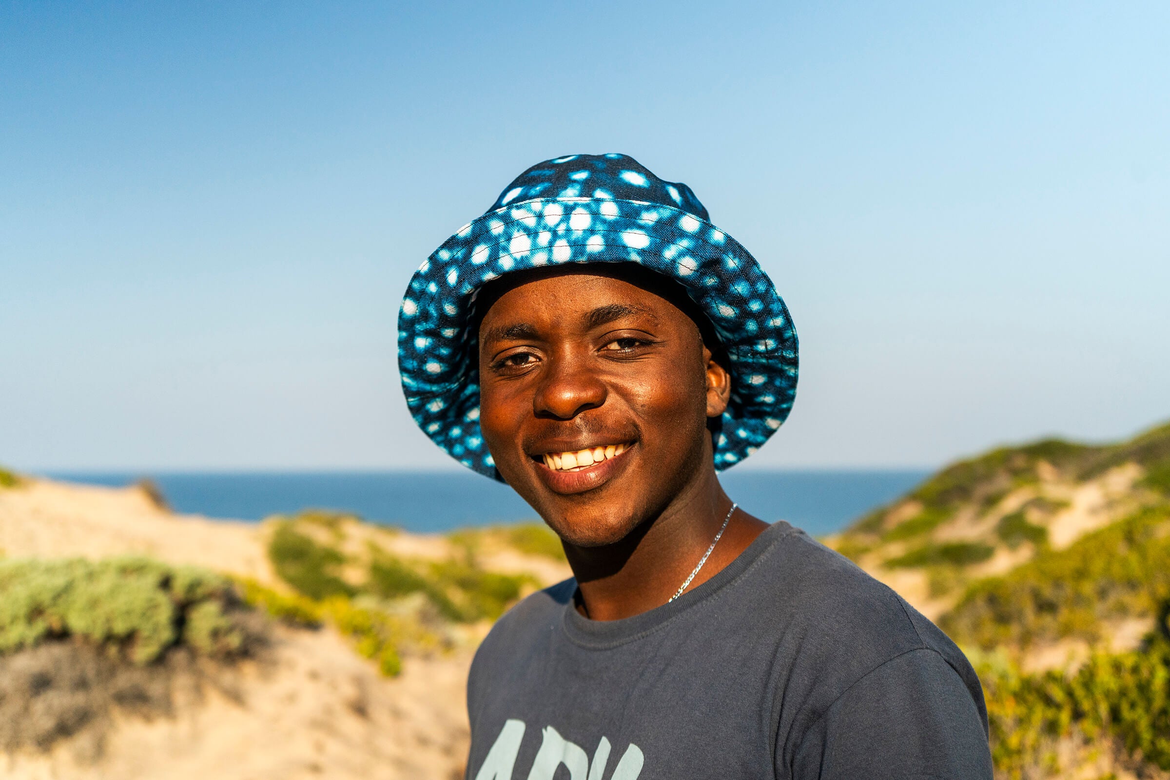Person wearing a patterned hat and gray shirt standing on a beach with ocean and sky in the background