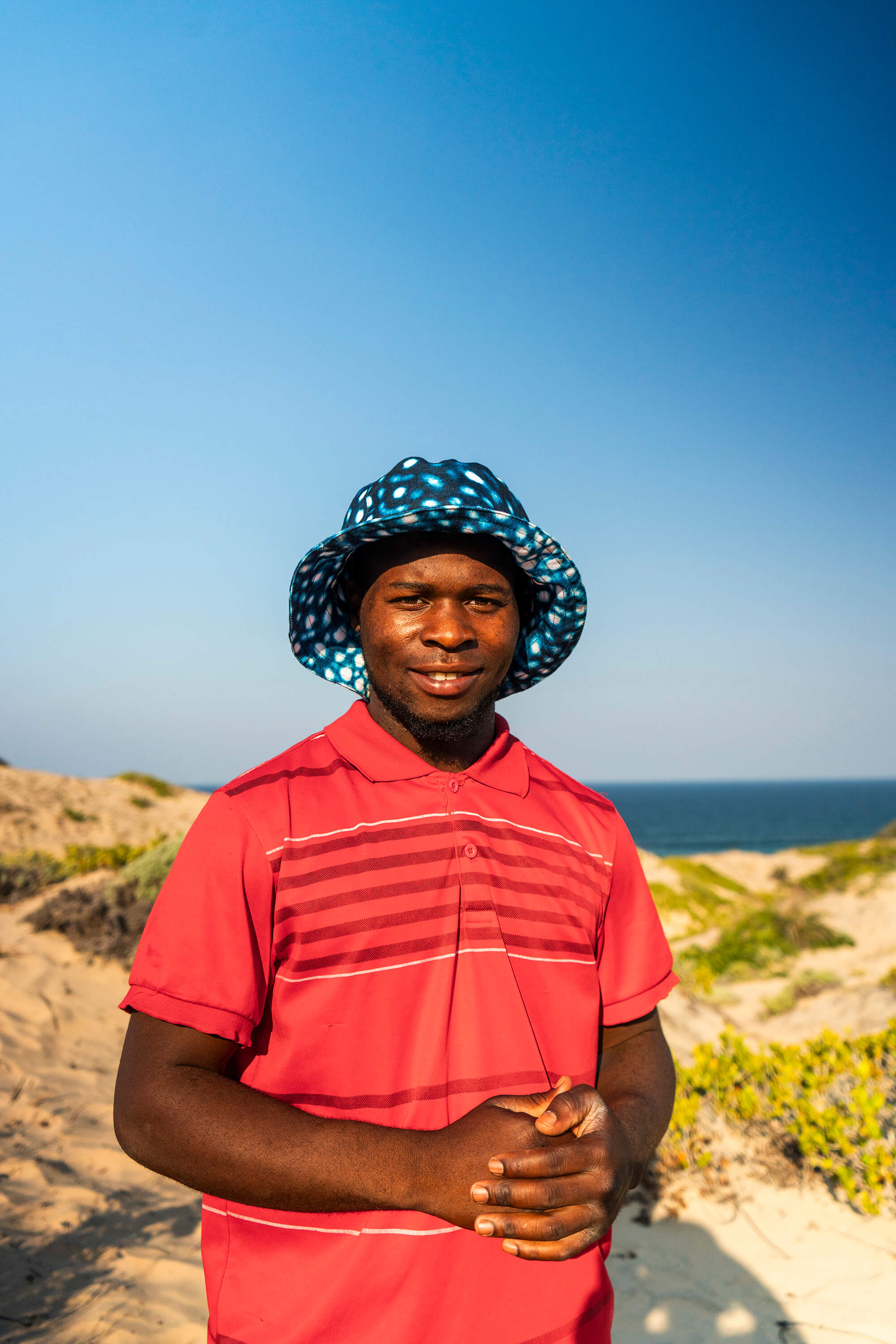 Person wearing a red shirt and patterned hat on a beach with blue sky and ocean.
